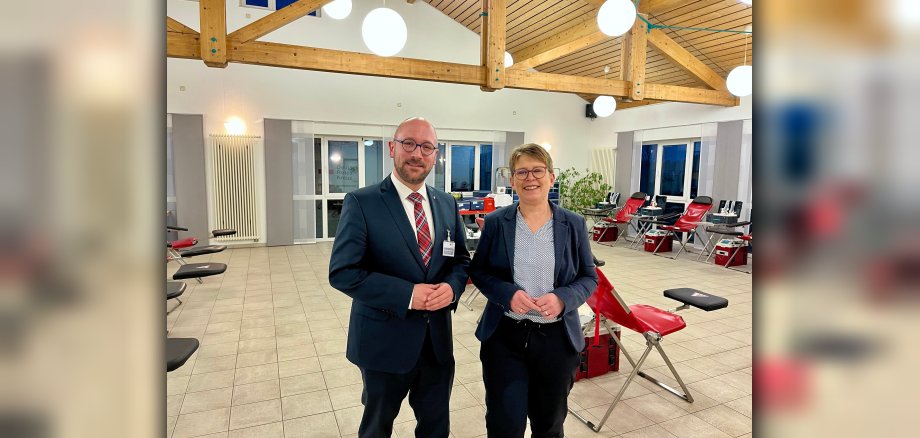 Das Foto zeigt Dr. Tanja Machalet und Benjamin Albrecht vor den leeren Plätzen, die zur Blutspende genutzt werden sollen. Es sind rote Stühle mit jeweils einer schwarzen Armstütze zu sehen.